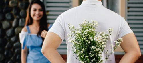 Mann steht vor Frau und versteckt Blumen für sie hinter seinem Rücken